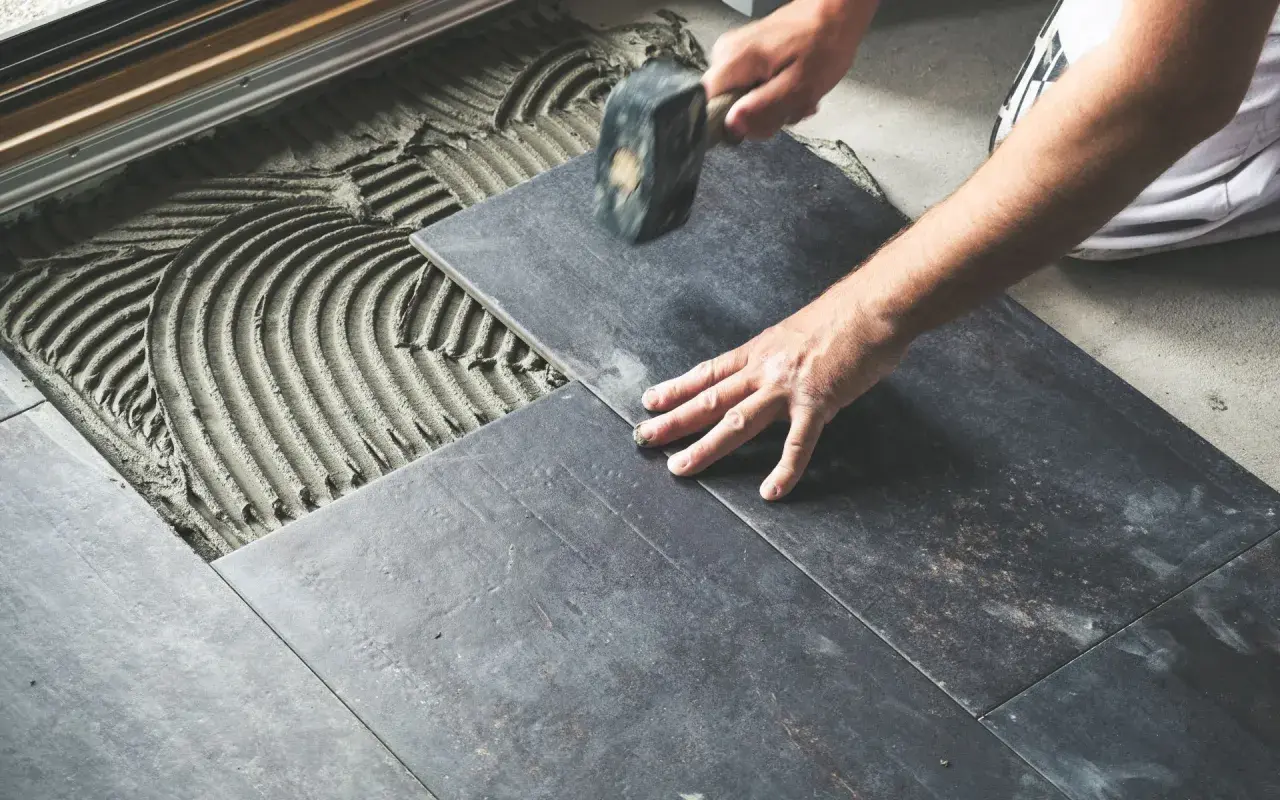 Mano che posa una piastrella scura su cemento fresco, con un martello per un posizionamento perfetto e rettificato.