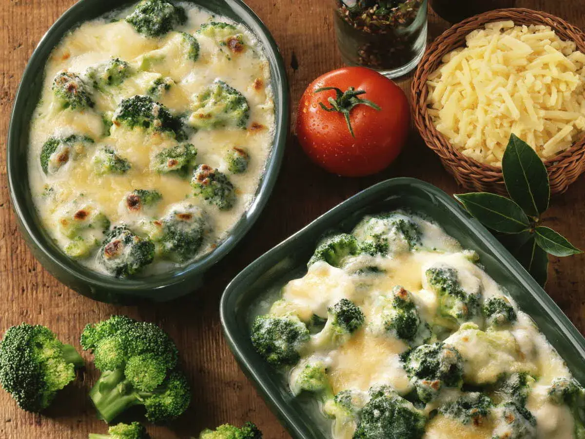 Deux plats de gratin de brocolis crémeux et gratinés, accompagnés de brocolis frais, d'une tomate et de fromage râpé.