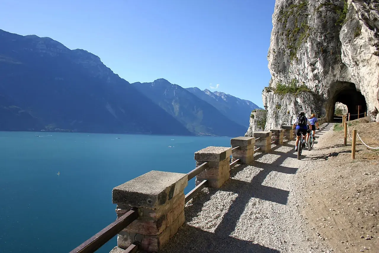 Ciclisti sul sentiero della Ponale, con il lago e le montagne sullo sfondo. Un tunnel scavato nella roccia attende.