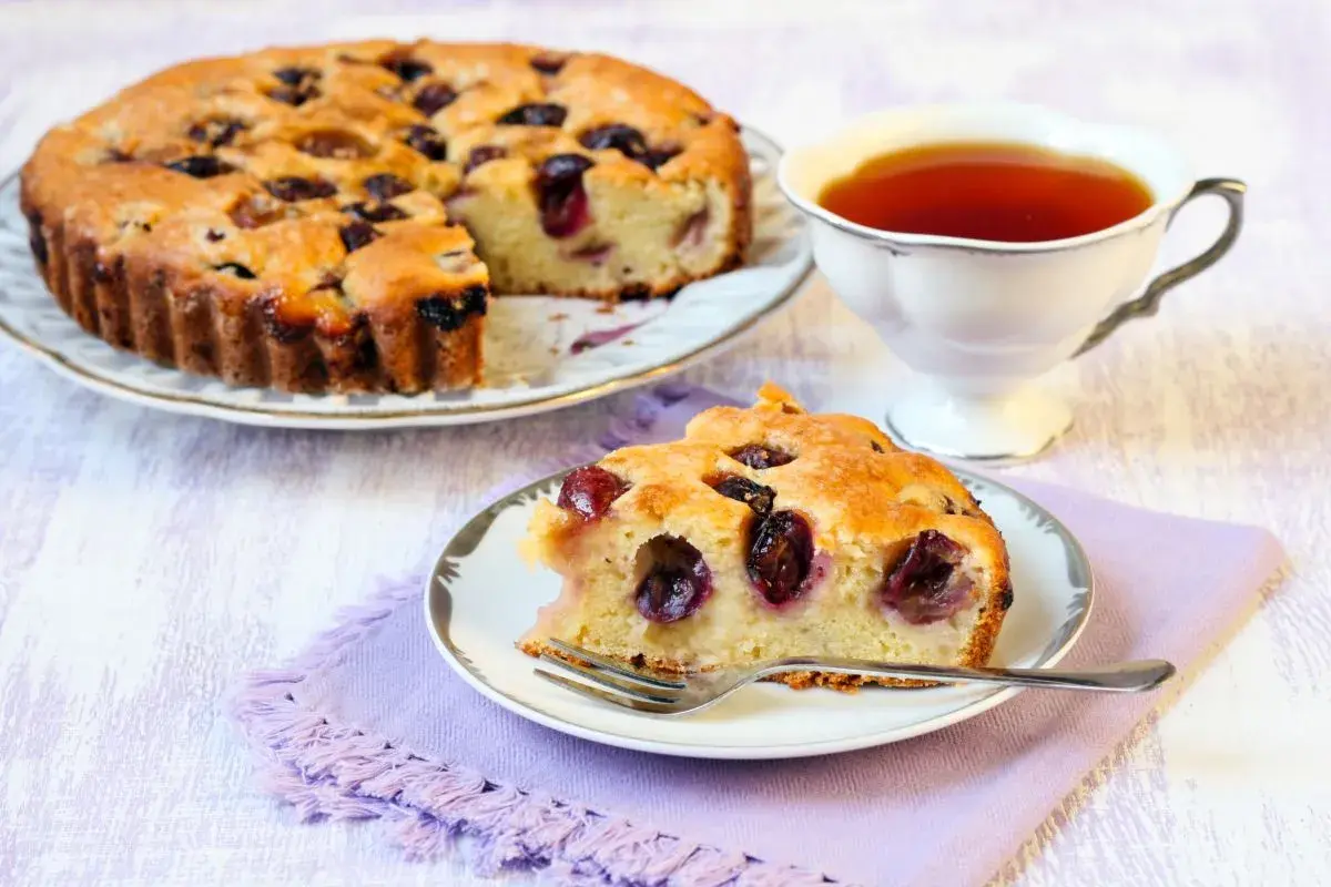 Una fetta di torta bertolina, soffice e profumata, con ciliegie mature, accanto a una tazza di tè fumante.