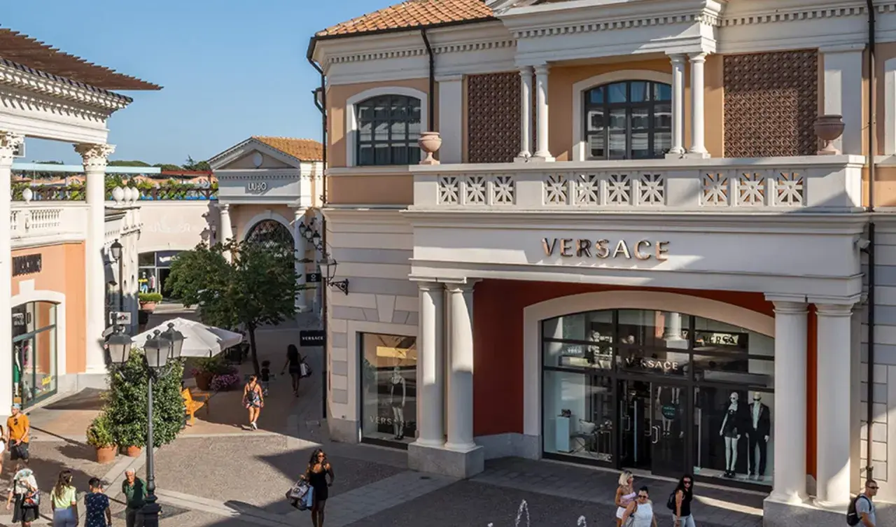 Boutiques de luxe, dont Versace, dans un centre commercial de style italien près de Rome. Des clients flânent sous le soleil.