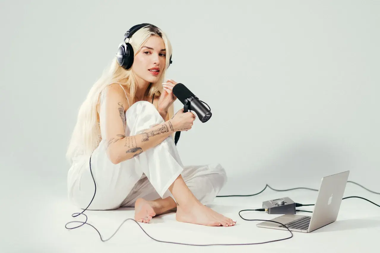 Elena Rose, a blonde woman with tattoos, sits on the floor wearing headphones and holding a microphone, ready to record.