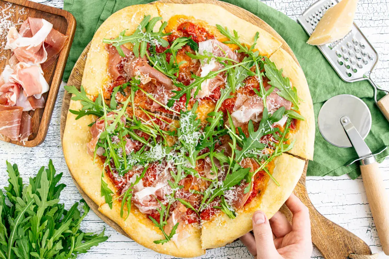 Pizza mit Rucola, Schinken und Parmesan, daneben Prosciutto, Parmesan und Pizzaschneider.