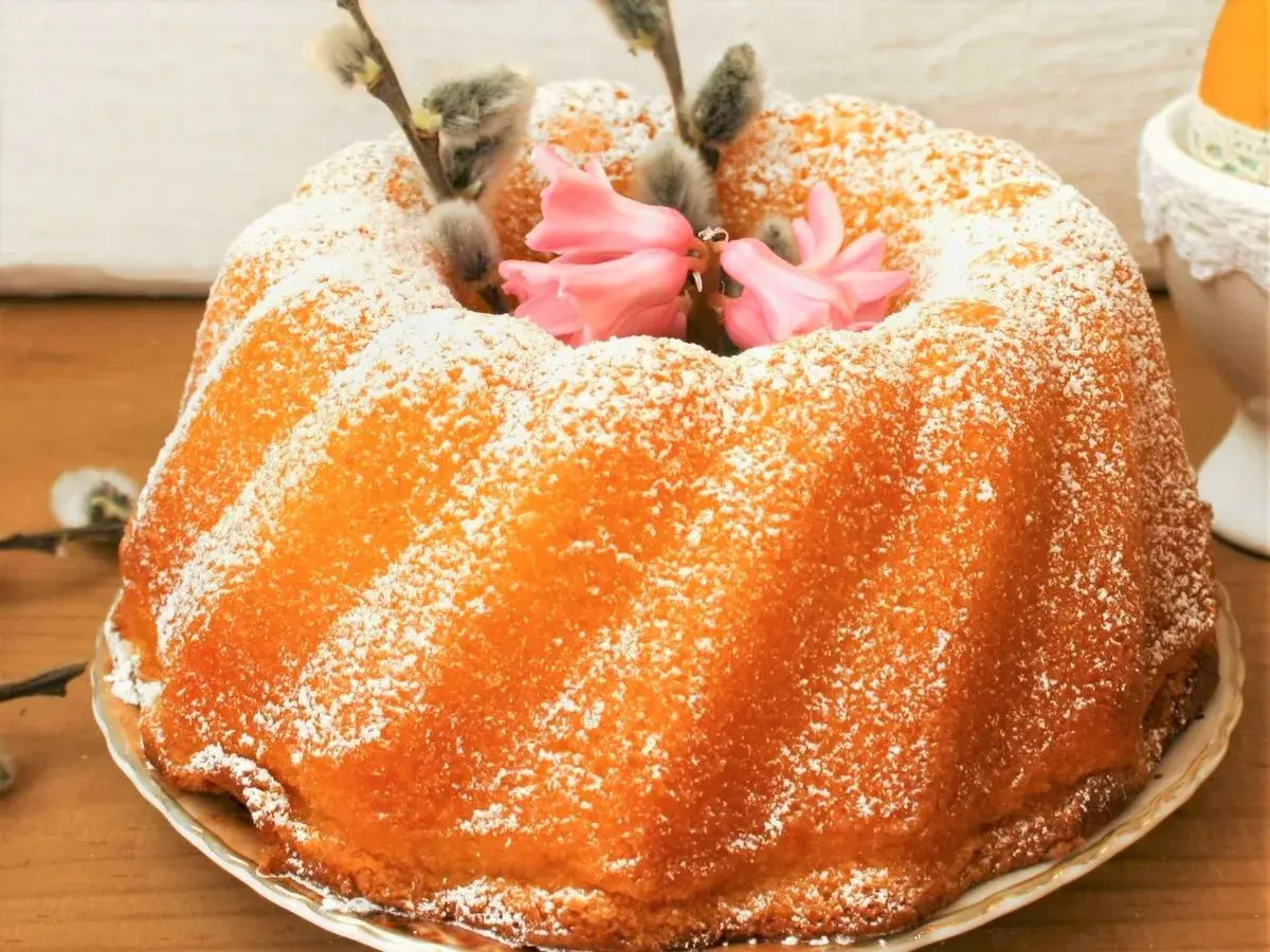 Puszysta babka jogurtowa oprószona cukrem pudrem, ozdobiona baziami i różowymi kwiatami. Idealna na wielkanocny stół.