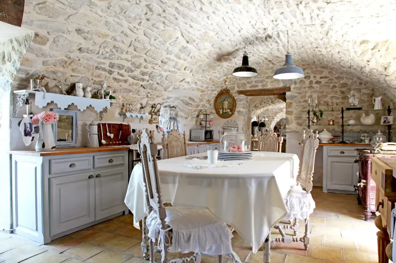 Cuisine rustique au style shabby chic, avec murs en pierre, table dressée et chaises anciennes.