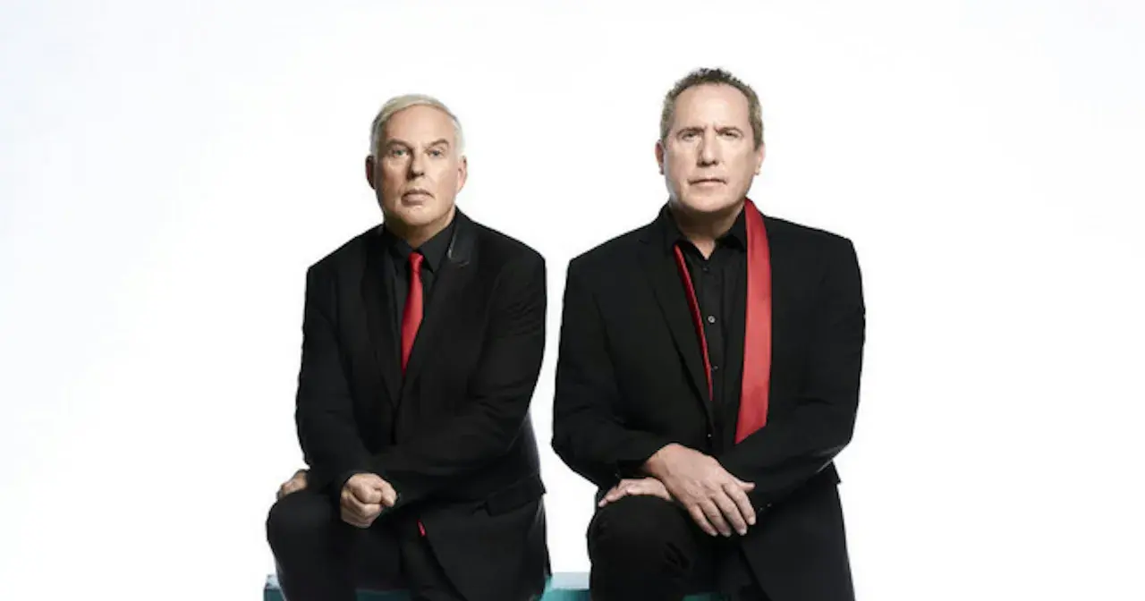 Andy McCluskey i Paul Humphreys z OMD, duet znany z niezapomnianych piosenek, w czarnych garniturach i czerwonych akcentach.
