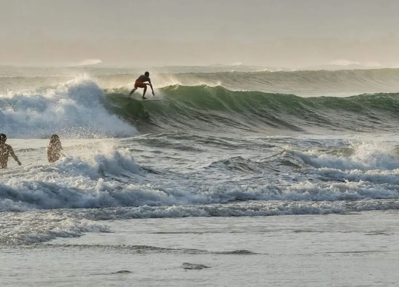Surfer na desce ślizga się po zielonej fali, podczas gdy dwie sylwetki stoją w wodzie. To dowód, że surfing w Polsce jest możliwy!