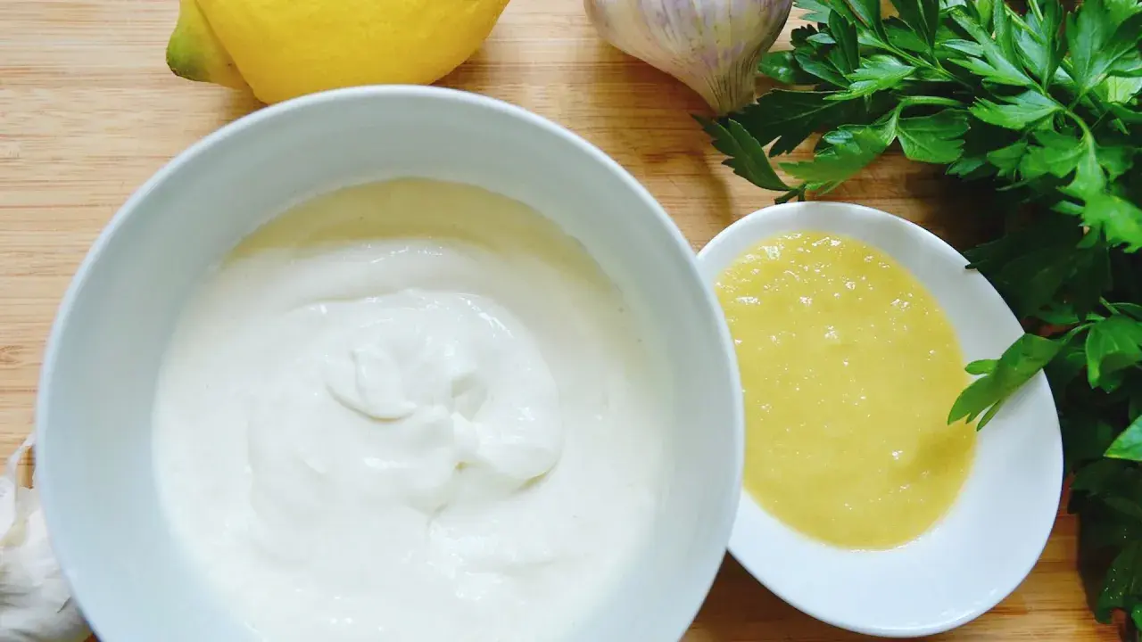 Zutaten für ein cremiges Aioli-Rezept: Knoblauch, Zitrone, Petersilie und zwei Schalen mit weißer und gelber Sauce.