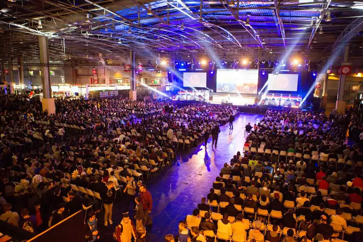 Gran multitud en un evento de convencion, sentada en sillas frente a pantallas gigantes con luces de escenario.