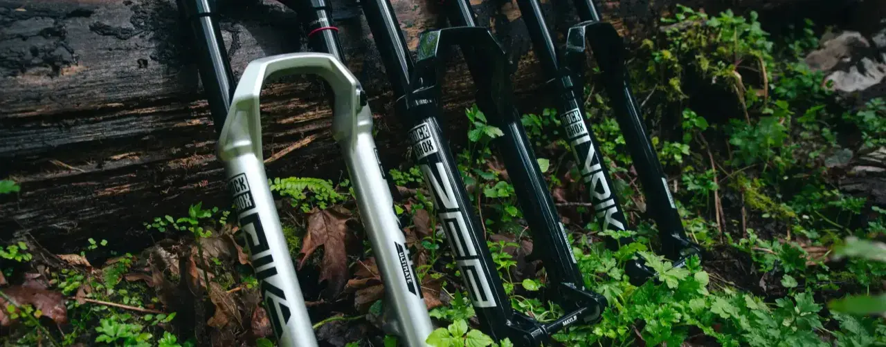 A silver RockShox Pike and black RockShox Lyrik forks lean against a log, ready for trail adventures.