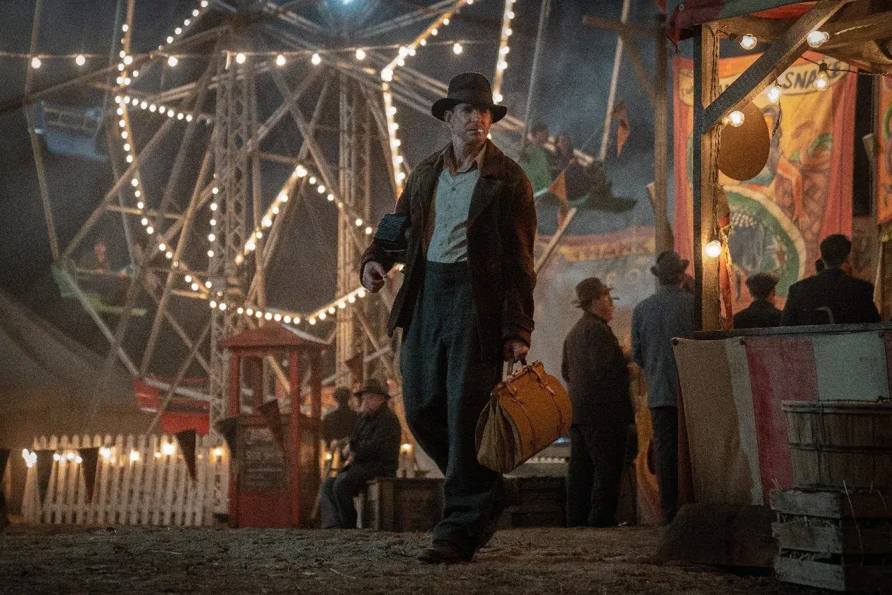 Un uomo con un cappello cammina in una fiera delle illusioni, con una ruota panoramica illuminata sullo sfondo. Le recensioni di Nightmare Alley descrivono questo mondo oscuro.