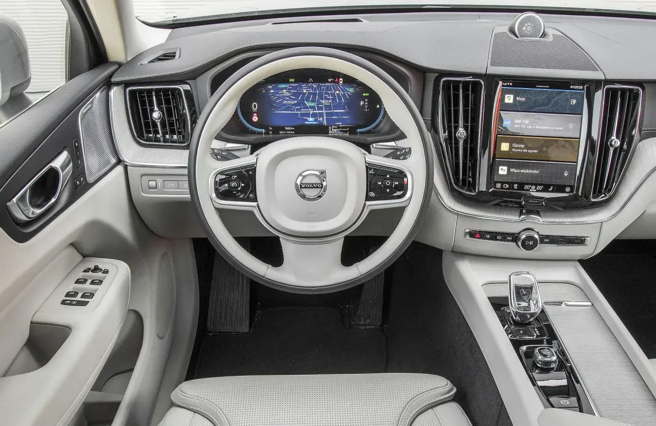 Wnętrze Volvo XC60 T6 z ekranem nawigacji i multimediów. Jasna tapicerka, biała kierownica z logo Volvo.