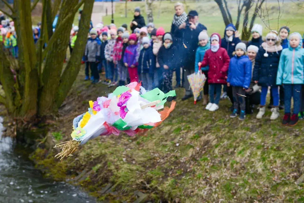 Dzieci obserwują topienie Marzanny, symboliczne pożegnanie zimy. Kolorowa kukła leci do wody.