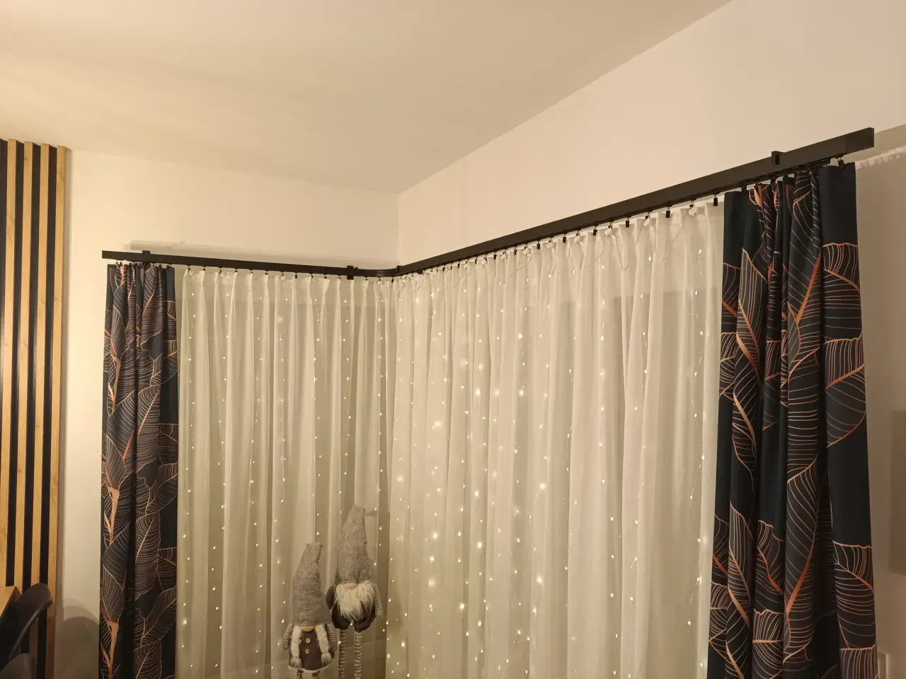 Elegancki karnisz apartamentowy czarny, na którym wiszą zasłony z motywem liści i delikatne firanki ze światełkami.
