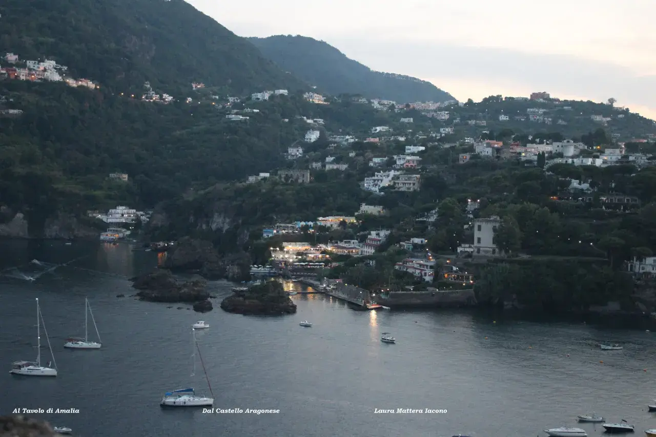 Vittoria Colonna a Ischia: case aggrappate alla collina, luci soffuse e barche a vela nella baia al crepuscolo.