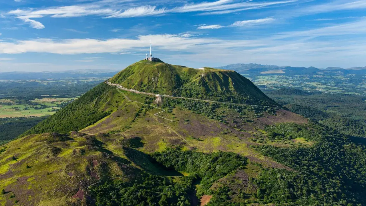 Vue aérienne du Puy de Dôme, un des volcans d'Auvergne, avec sa route en lacets et son antenne. Paysage verdoyant sous un ciel bleu.