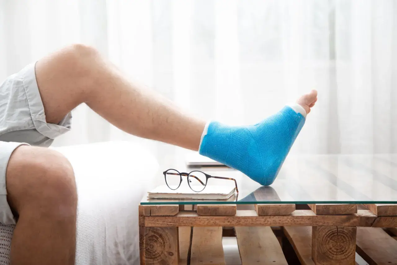 Mann mit blauem Gipsbein ruht sich aus. Die Frage "welche schuhe nach fersenbeinbruch" beschäftigt ihn, während er auf einem Holztisch mit Glasplatte entspannt.