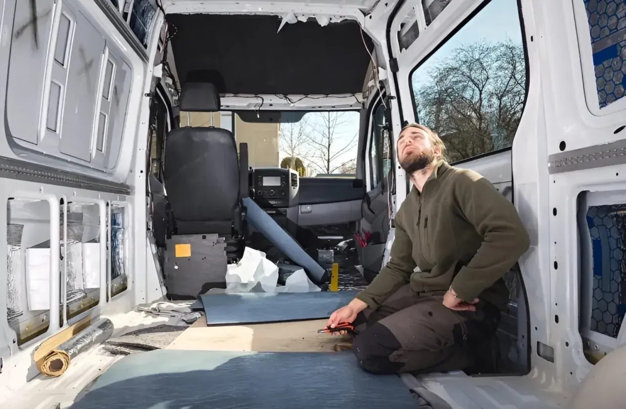 Uomo lavora all'isolamento di un'auto camperizzata. Materiali sparsi, sedili rimossi, pronto per la trasformazione.