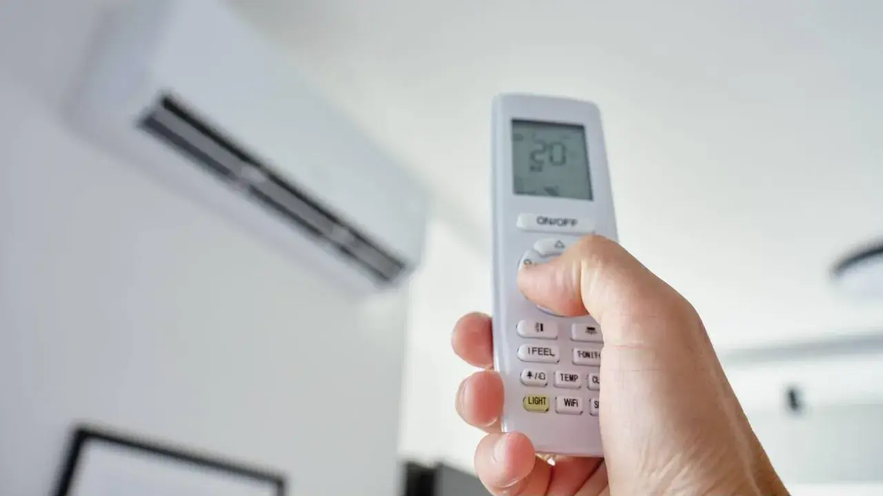 Mano sujetando un control remoto para encender aire acondicionado. La unidad interior está en el fondo.