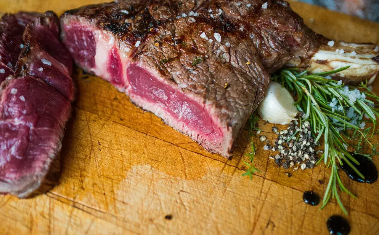 Une belle côte de bœuf saignante, assaisonnée de gros sel et de poivre, avec du romarin et de l'ail sur une planche en bois.