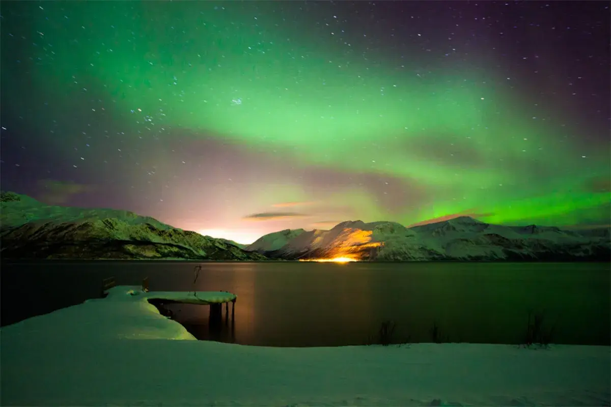 Spektakuläre Nordlichter über schneebedeckten Bergen und einem gefrorenen See. Ein perfekter Winterurlaub in Skandinavien.