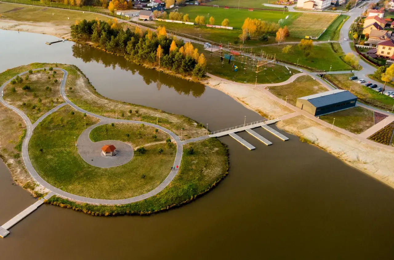 Malownicze, nisko jezioro z wyspą, ścieżkami, altanką i nowoczesnym budynkiem. W tle widać drzewa, plażę i zabudowania.