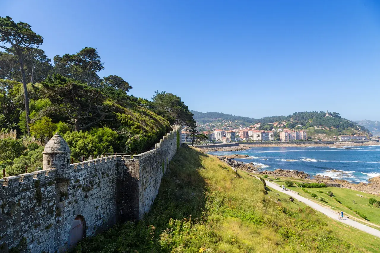 A historic stone wall winds along the coast of Baiona, Portugal, overlooking the blue sea and a town nestled on the hills.