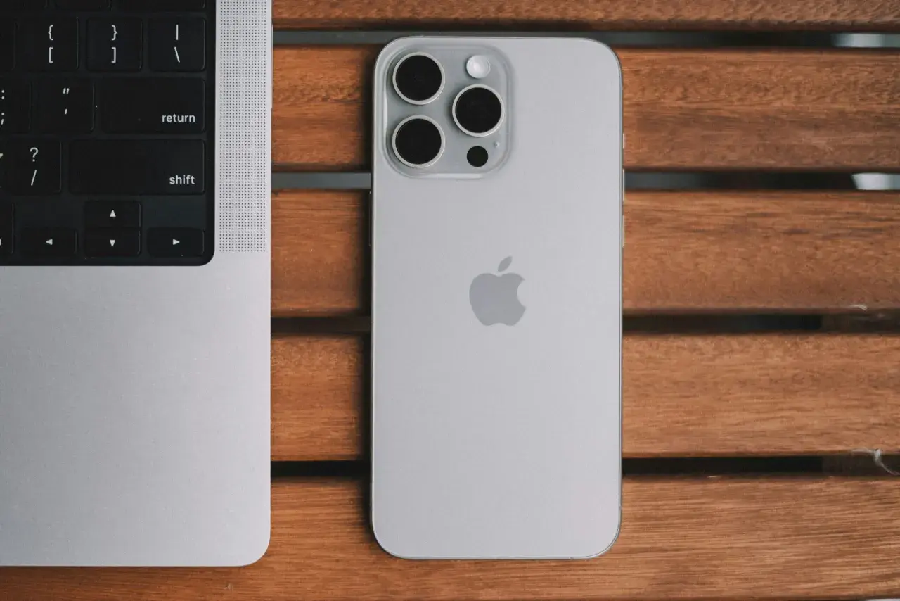 Srebrny iPhone 13 Pro z systemem iOS 26.4 obok klawiatury MacBook'a na drewnianym stole.