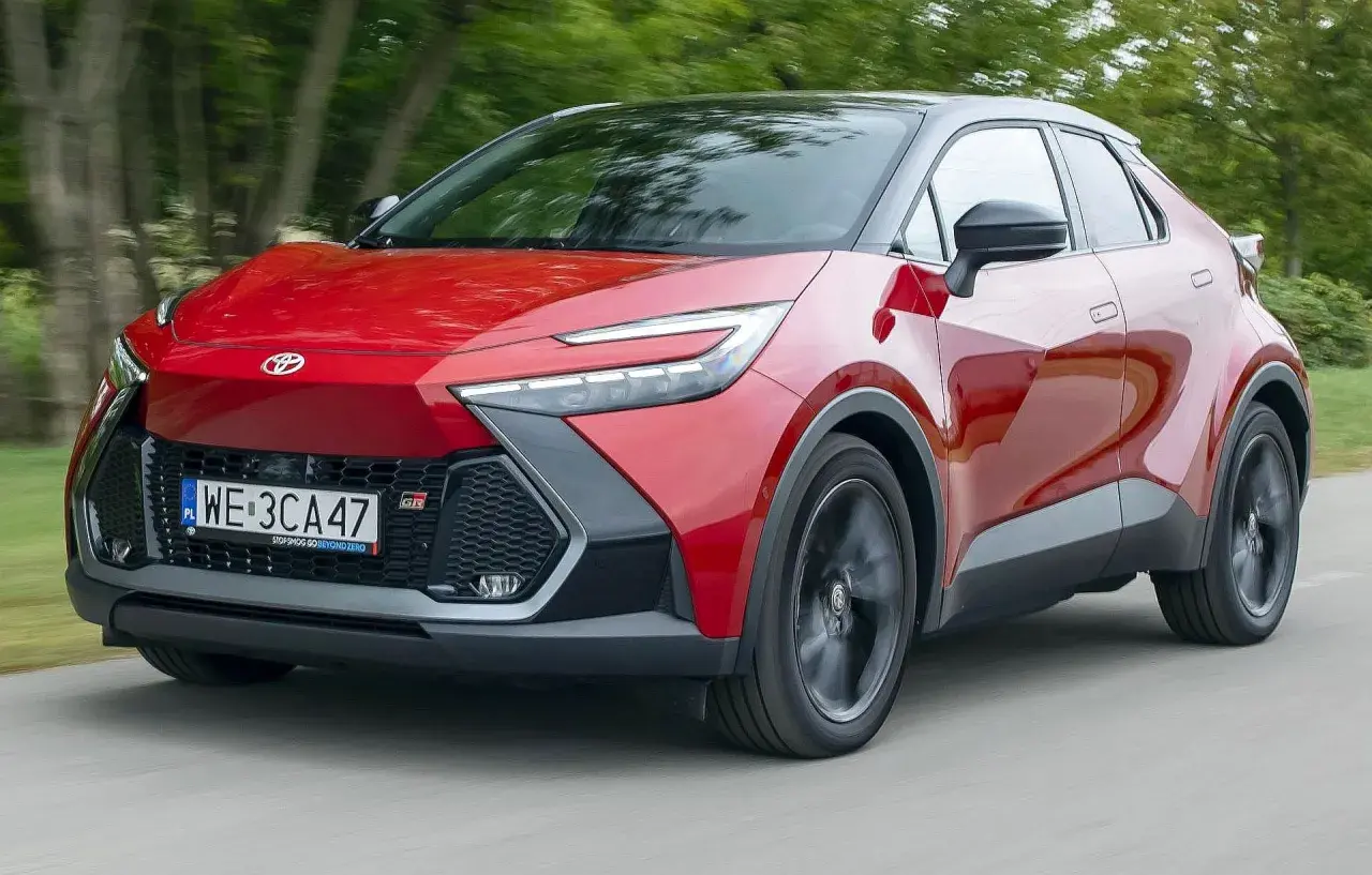 Czerwona Toyota C-HR 2020 z czarnym dachem pędzi po drodze, otoczona zielenią drzew.