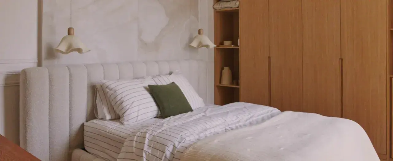 Una lámpara mesilla de madera ilumina una cama con ropa de cama a rayas y un cabecero texturizado, junto a un armario de madera.