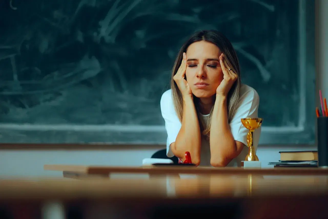 Lehrerin mit Händen an den Schläfen, Augen geschlossen, wirkt erschöpft. Ein Pokal und Bücher auf dem Tisch deuten auf den Schulalltag hin, der zu Burnout bei Lehrern führen kann.