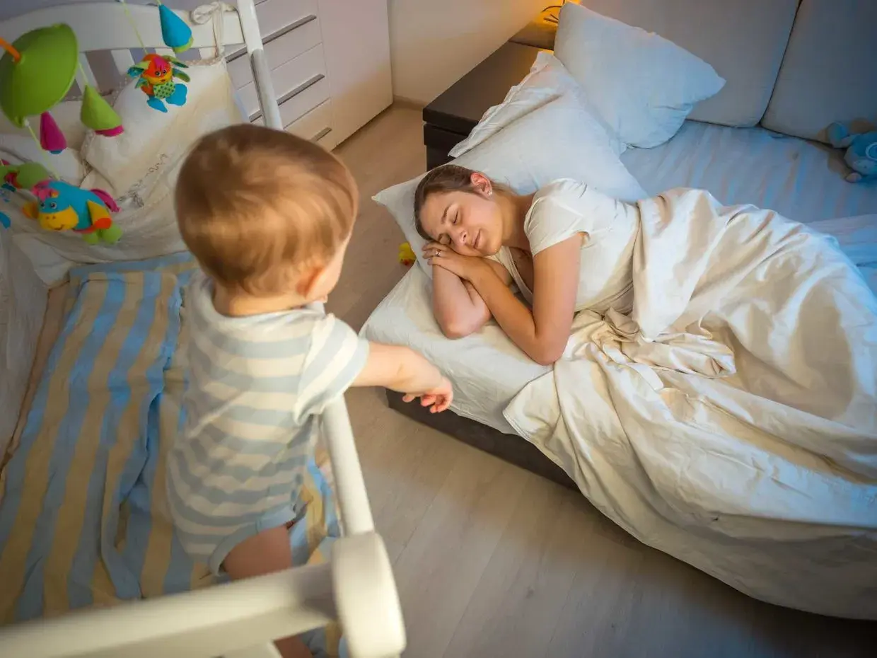 Un neonato di 7 mesi, in piedi nella sua culla, osserva la mamma addormentata nel letto.