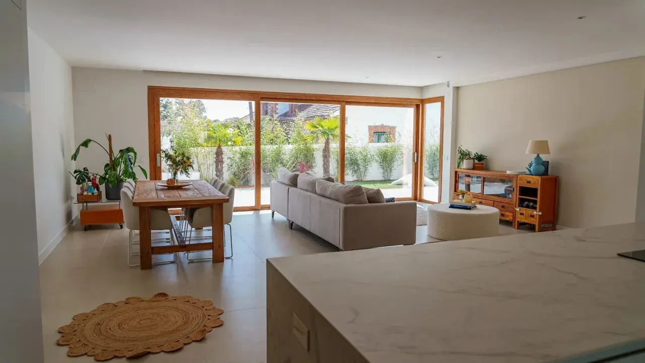 Salón moderno con comedor y sofá, destacando una pared prefabricada interior que se integra con grandes ventanales al jardín.