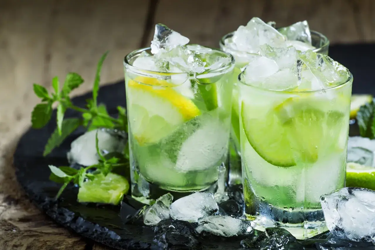 Trois verres de caipirinha glacée, garnie de citron vert et de citron, sur un plateau noir avec de la menthe fraîche. La recette parfaite pour se rafraîchir.