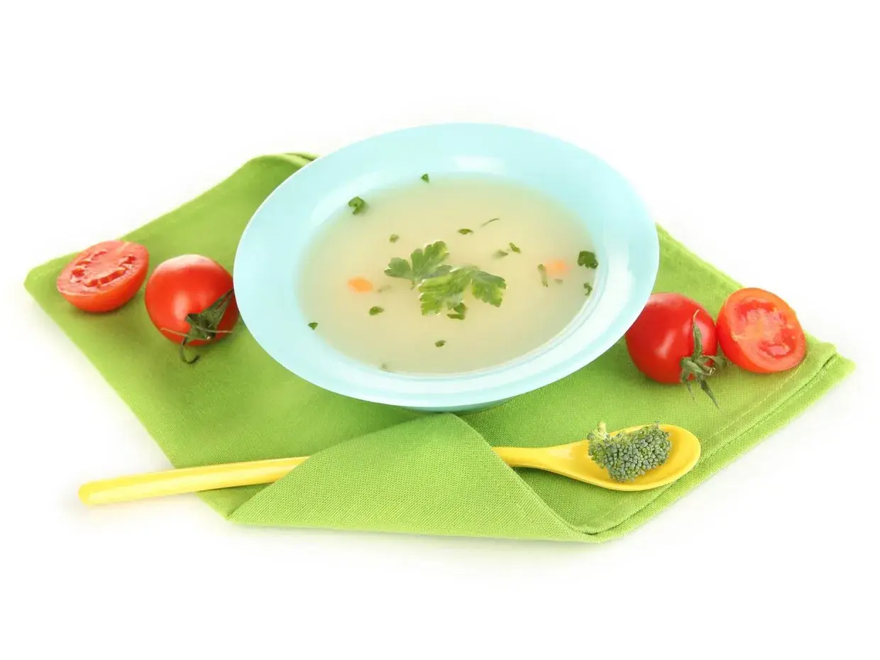 Brodo vegetale neonati in ciotola azzurra, con pomodorini e un cucchiaino giallo con broccoli.