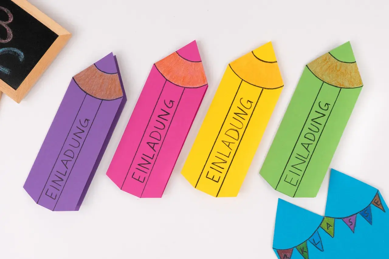 Bunte Stift-Bastelvorlagen mit "Einladung" beschriftet. Eine kleine Tafel mit ABC und eine blaue Wimpelkette sind ebenfalls zu sehen.