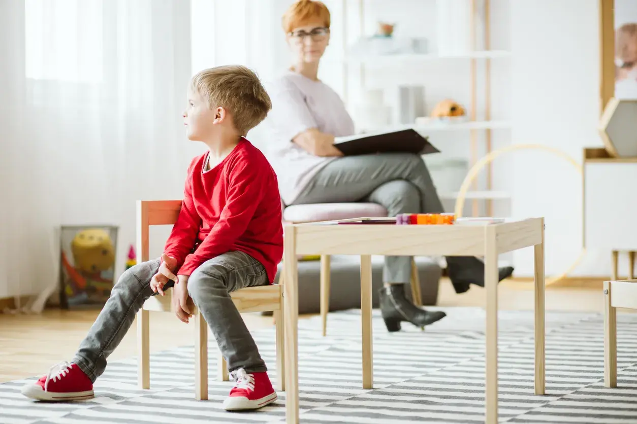 Pojke i röd tröja sitter på en stol under en autism utredning barn. En kvinna sitter bakom ett bord.
