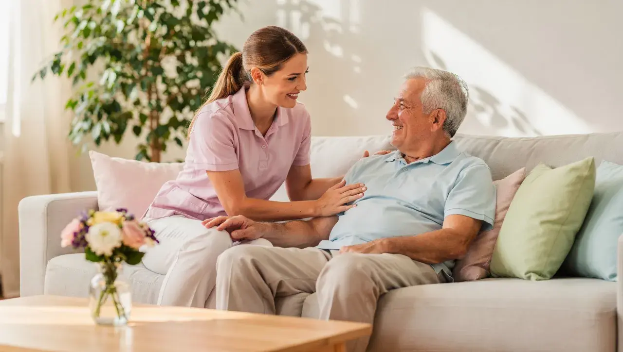 Eine Pflegerin spricht lächelnd mit einem älteren Mann auf einem Sofa. Ein Gespräch über den Eigenanteil im Pflegeheim kann so angenehm sein.
