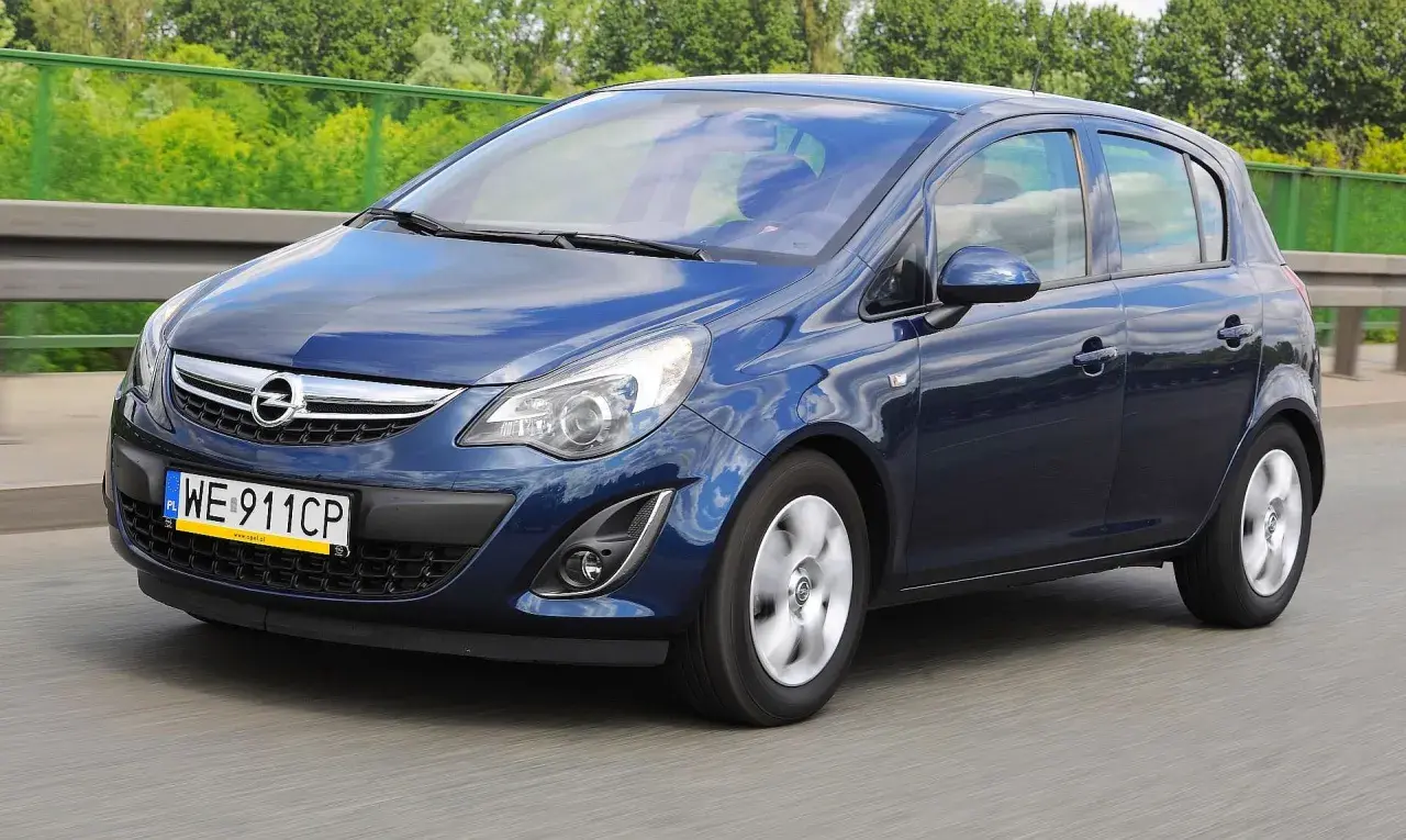 Granatowy Opel Corsa D jedzie po drodze, odbijając niebo i chmury.
