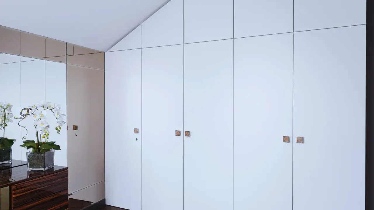 Armario empotrado con puertas blancas y tiradores cuadrados. Reflejos de orquídeas y un mueble de madera oscura en el espejo. Ideal para cambiar puertas armario empotrado.