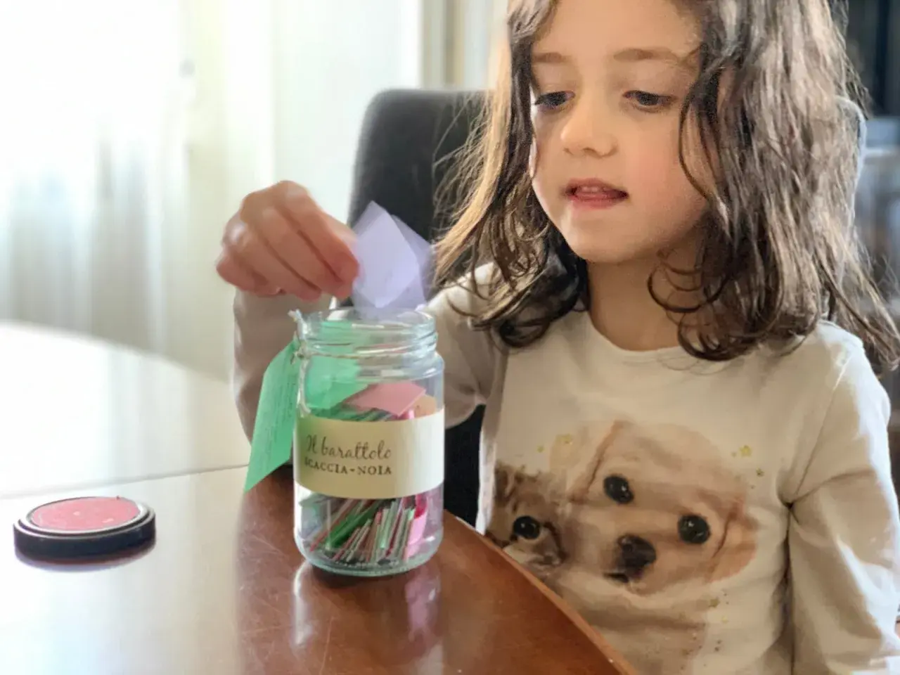 Bambina inserisce un bigliettino nel barattolo "Scaccia-Noia", pieno di idee per giochi da fare quando ci si annoia.