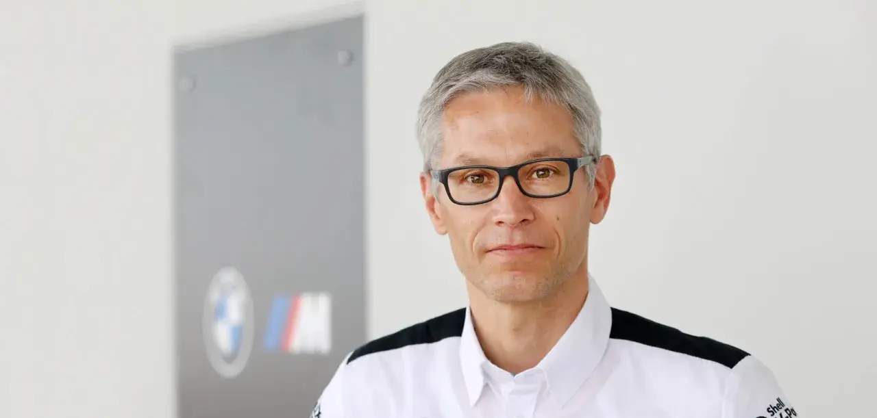 Mike Krack, con gafas y camisa blanca con detalles negros, posa frente a un logo de BMW M.