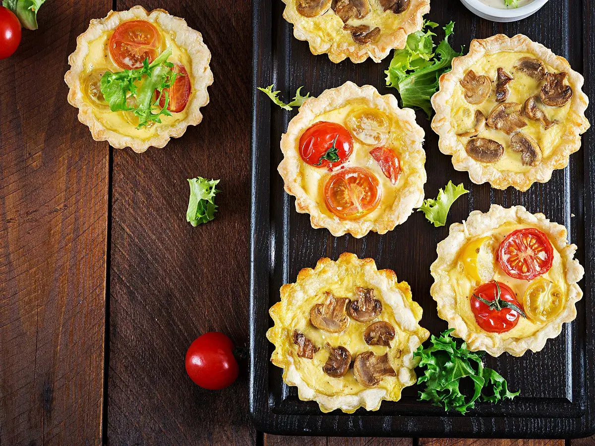 Mini quiches apéro aux tomates et champignons, garnies de feuilles de salade verte, sur fond de bois sombre.