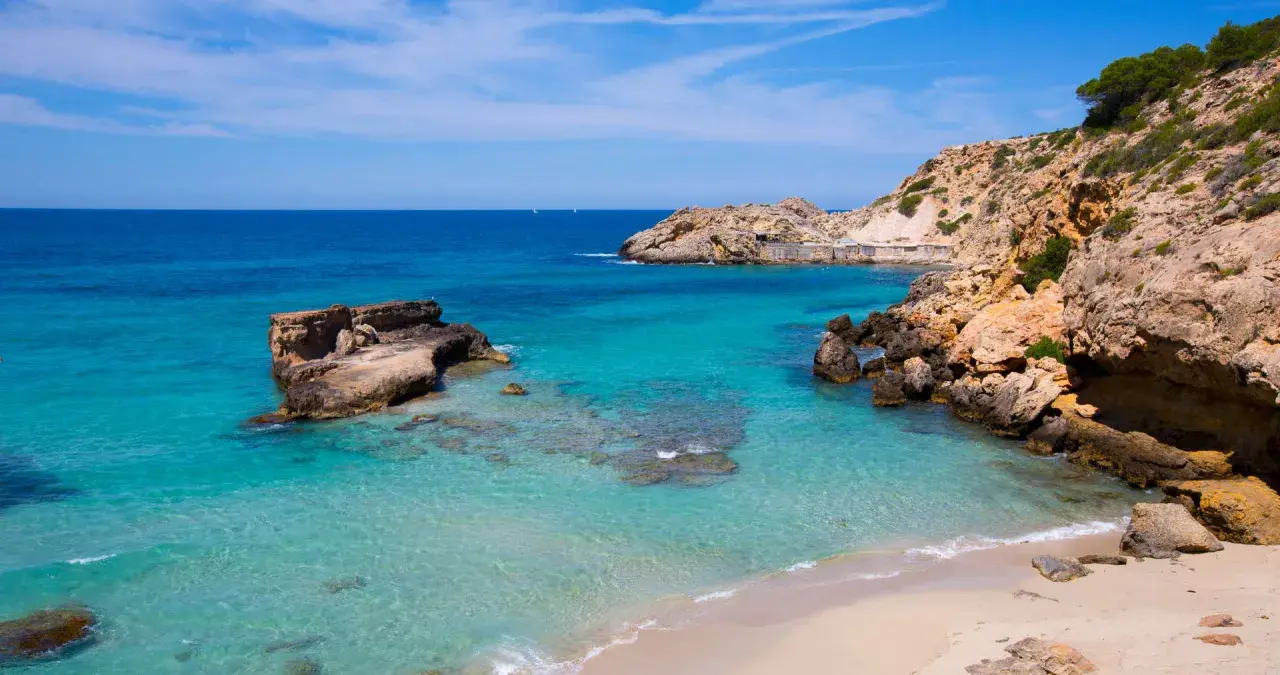 Aguas cristalinas y rocas escarpadas en una cala idílica. Unos pequeños **planes** se ven en el horizonte, invitando a soñar con esta **playa** paradisíaca.