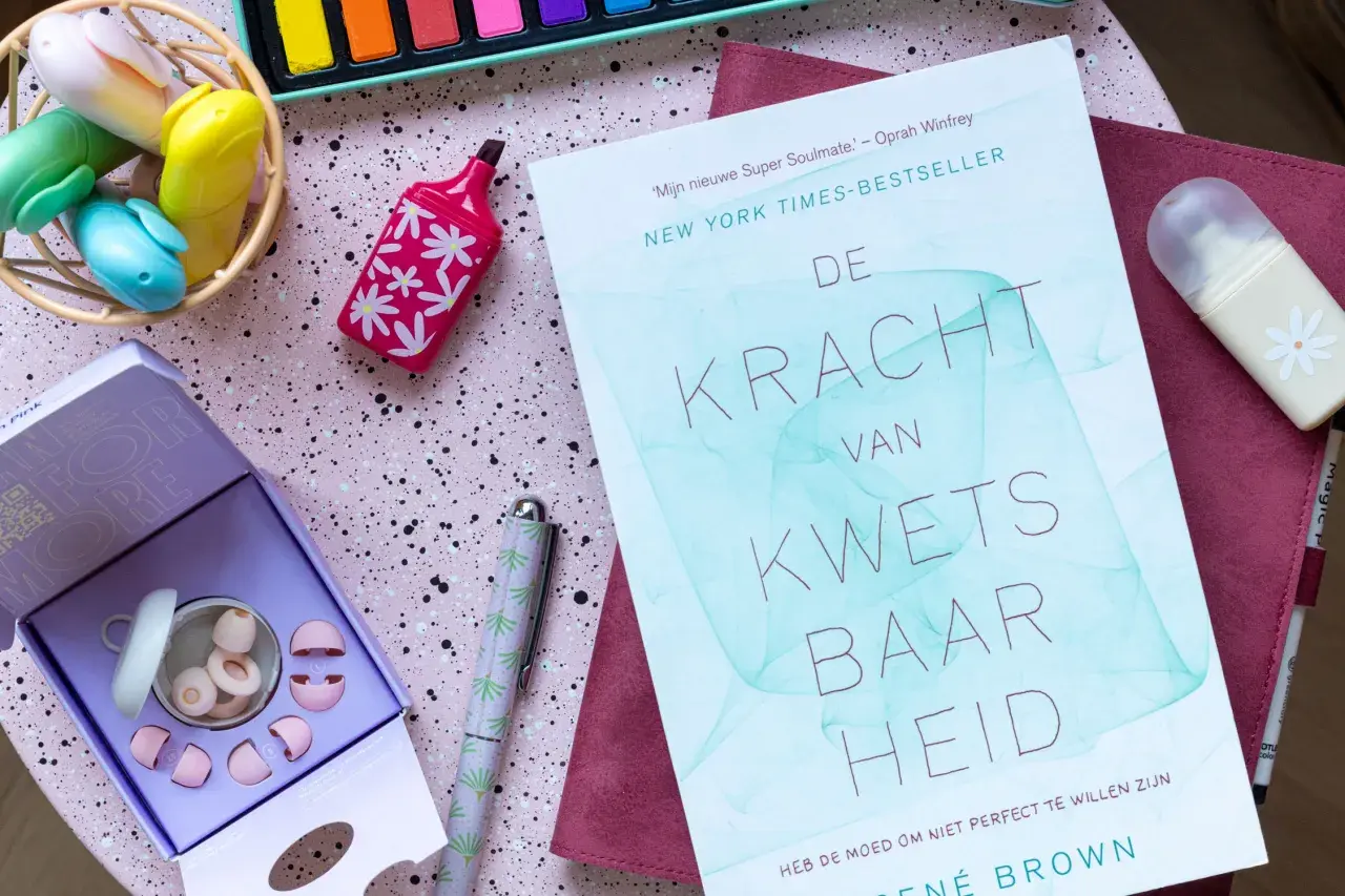 Boek 'De Kracht van Kwetsbaarheid' en oordopjes, ideaal voor wie last heeft van omgevingsgeluid. Creatieve spullen op een roze ondergrond.