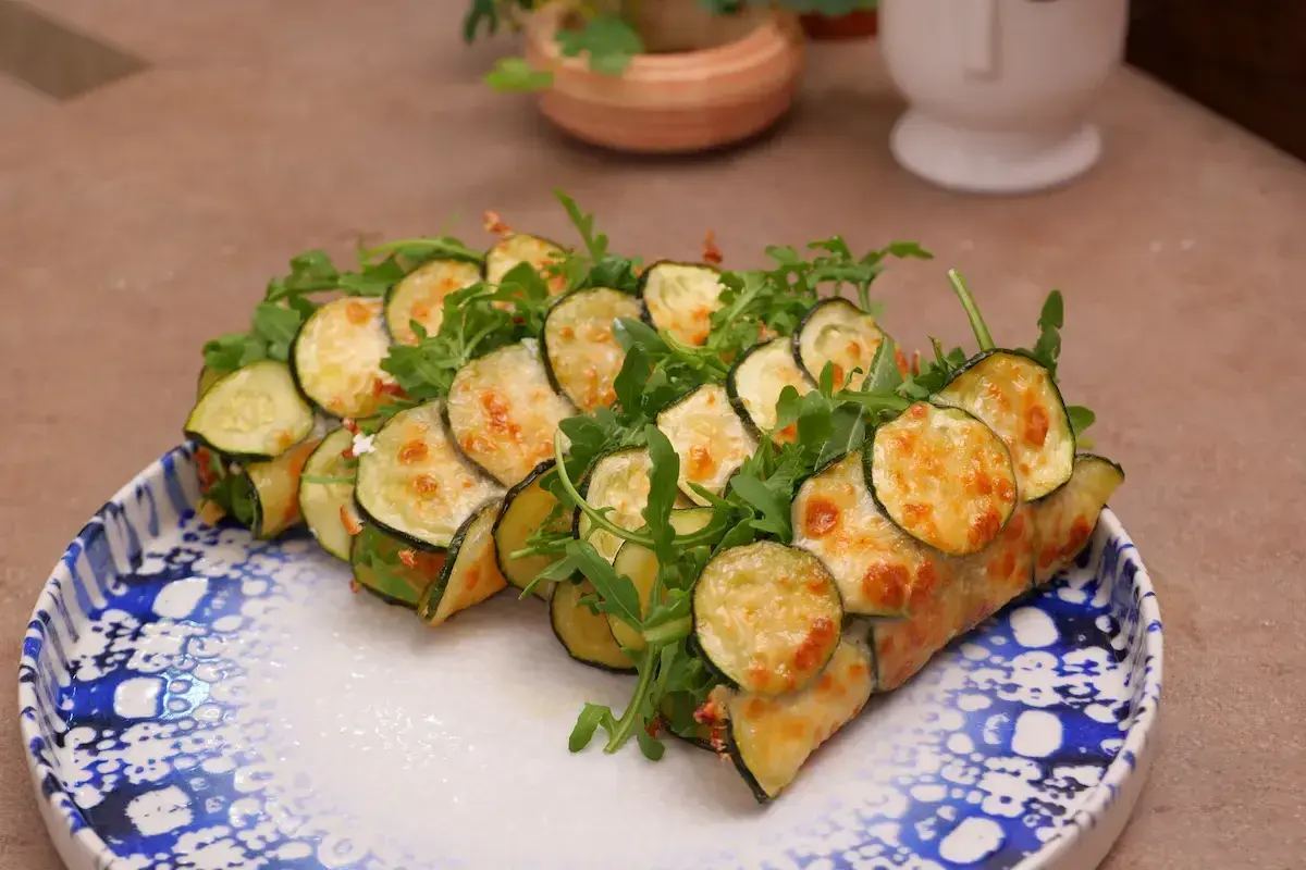 Rotolini di zucchine e rucola, cotti in padella e gratinati con formaggio fuso, serviti su un piatto decorato.