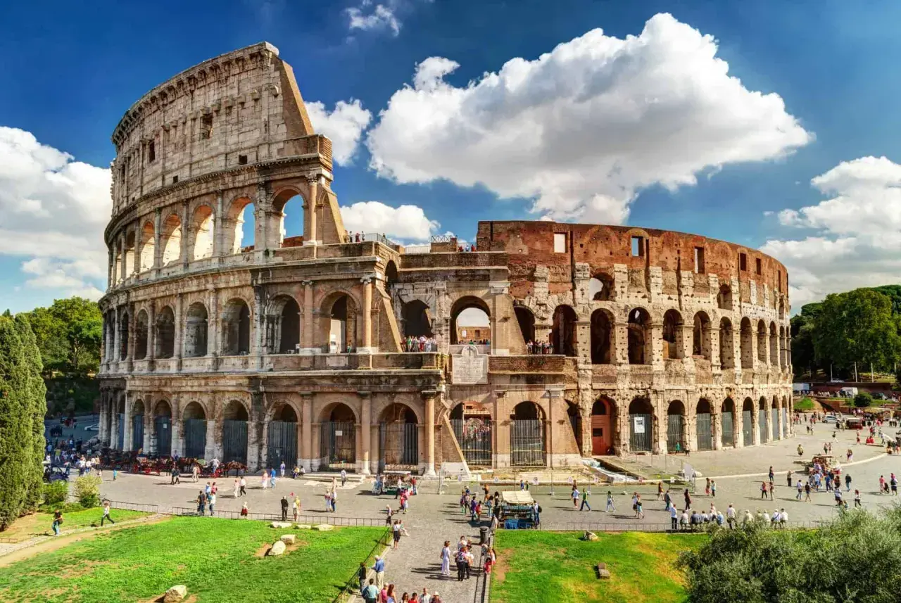 Le Colisée de Rome en août, baigné de soleil, accueille des visiteurs sous un ciel bleu parsemé de nuages.