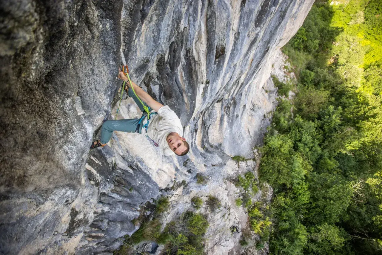 Un grimpeur escalade une paroi rocheuse, concentré sur sa prochaine prise. La végétation luxuriante contraste avec la pierre grise.