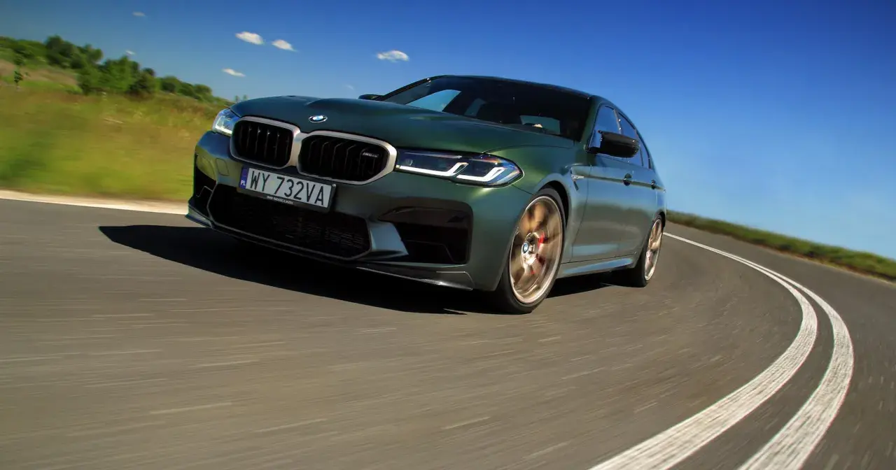 Zielony BMW M5 CS na zakręcie drogi, z rozmytym tłem i niebieskim niebem.