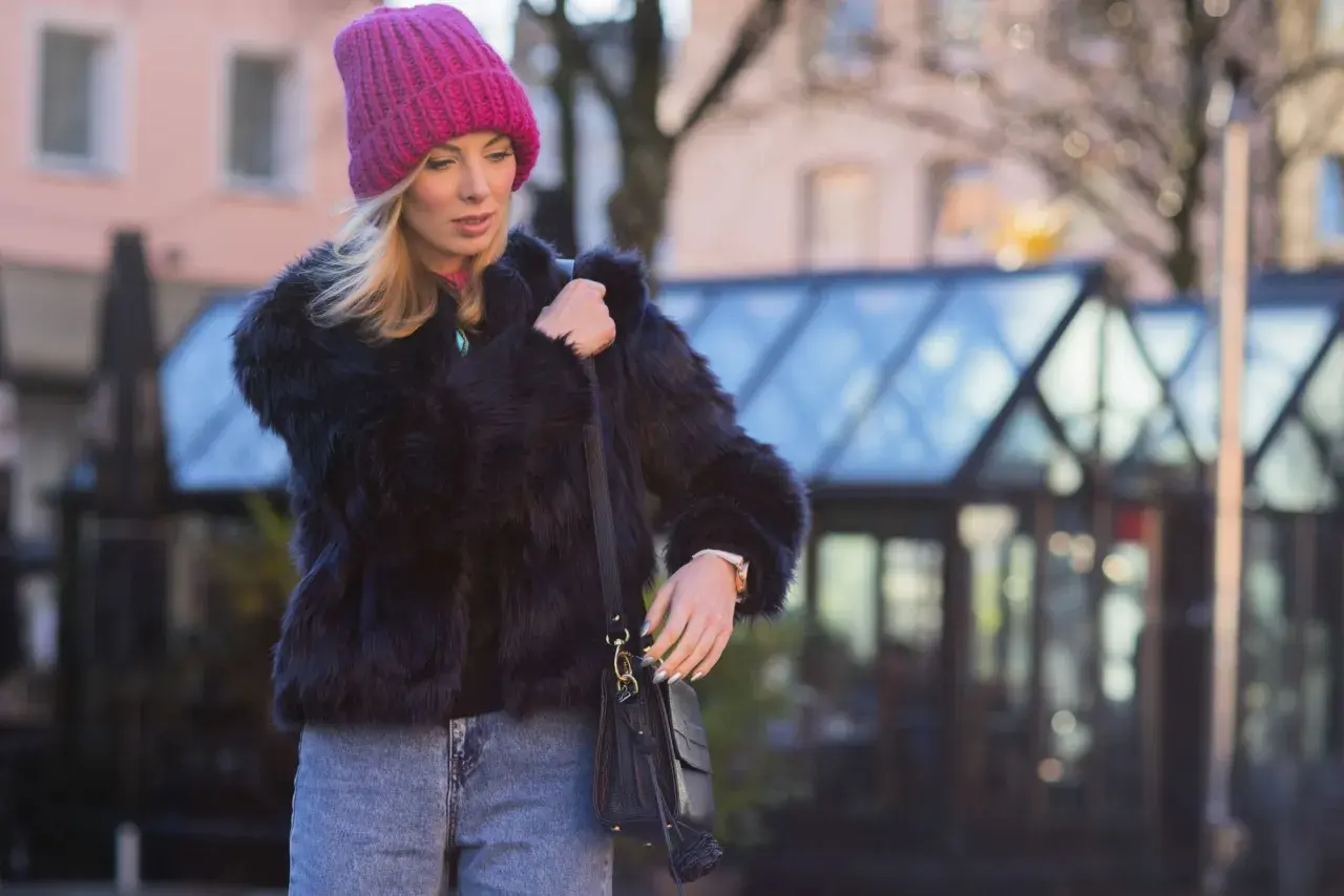 Krótkie futerko stylizacje: blondynka w różowej czapce i jeansach, z czarną torebką.